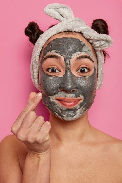 Close up shot of beautiful Asian woman applies purifying black mask on face, has beauty treatments, makes korean like sign, wears grey headband, stands shirtless against pink wall in bathroom