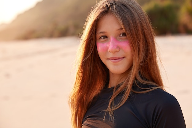 Close up shot of beautiful Caucasian girl with pink surf zinc mask, dressed in black wetsuit