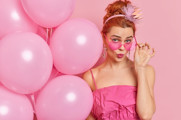 Close up shot of beautiful charming redhead glamour young woman looks from under trendy pink sunglasses dressed in fashionable outfit holds helium balloons stands indoor celebrates birthday.