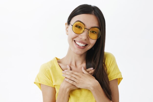 Close-up shot of beautiful charming woman with no makeup, having acne standing in yellow round sunglasses holding palms on chest