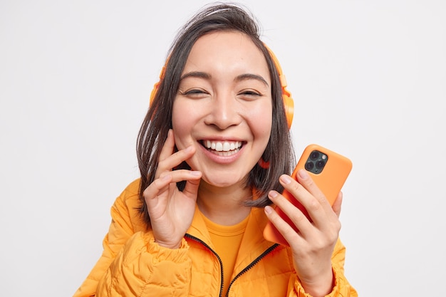 Close up shot of beautiful cheerful Asian woman smiles broadly from delight enjoys listening favorite music holds mobile phone dressed in orange jacket isolated over white  wall.