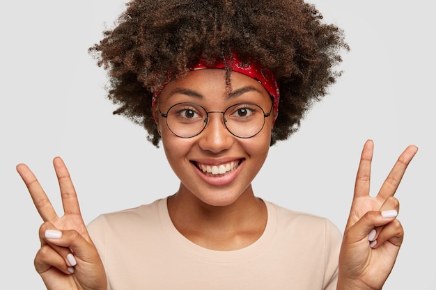 Close up shot of beautiful cheerful dark skinned young woman with curly hair makes peace sign with both hands