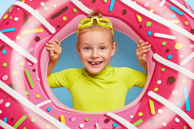 Close up shot of beautiful cheerful redhead little girl wears swim goggles on head, looks through pink rubber swimring, has broad smile, lack of teeth, enjoys last days of hot summer, sunny day