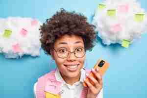 Free photo close up shot of beautiful curious female office worker smiles happily holds mobile phone checks newsfeed surrounded by colorful stickers with written information or list to do