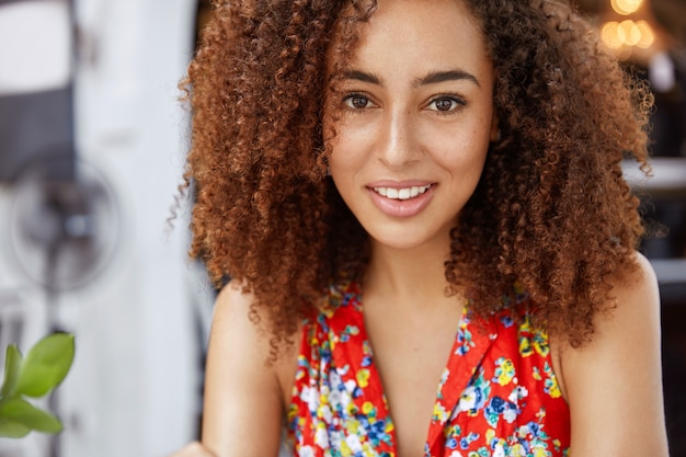 Free photo close up shot of beautiful dark skinned curly woman looks with pleased expression, comes on meeting with colleague to discuss collaboration