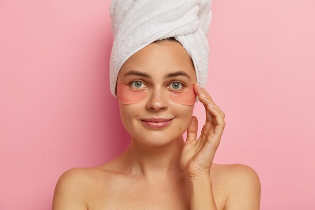 Close up shot of beautiful European female model makes spa procedures after taking shower, applies collagen patches under eyes, has anti aging treatment, stands indoor