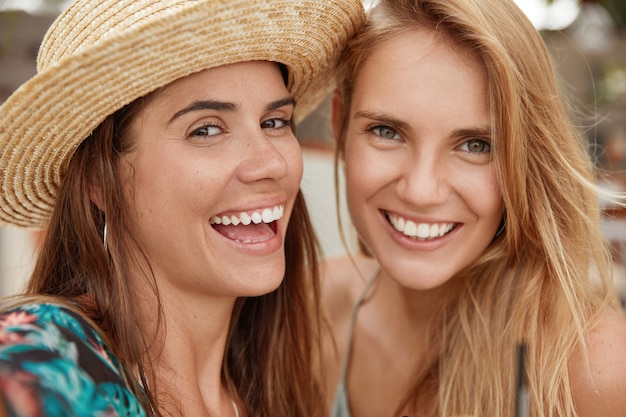 Close up shot of beautiful females with broad smiles, have positive expression, happy to meet together . Smiling young brunette woman in straw hat spends summer vacations with friend