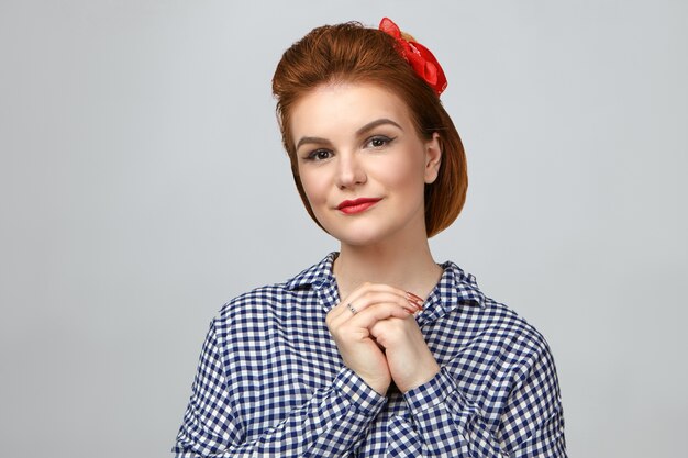 Close up shot of beautiful gorgeous young woman wearing retro hairdo, red lipstick and plaid shirt, looking at camera with calm smile, holding hands together. Beauty, style and fashion concept