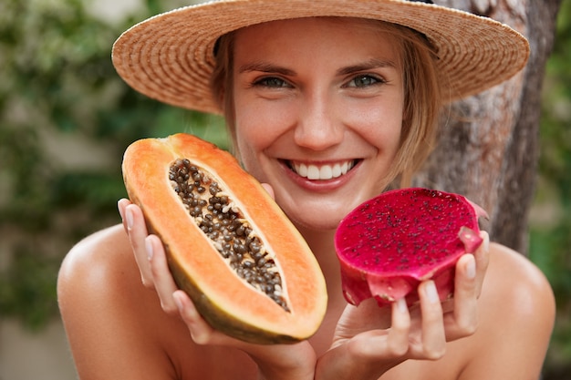 Close up shot of beautiful smiling female with appealing appearance, pleasant smile, holds papaya and dragon fruit, poses outdoor in tropical place, eats juicy delicious fruits. summer trip.