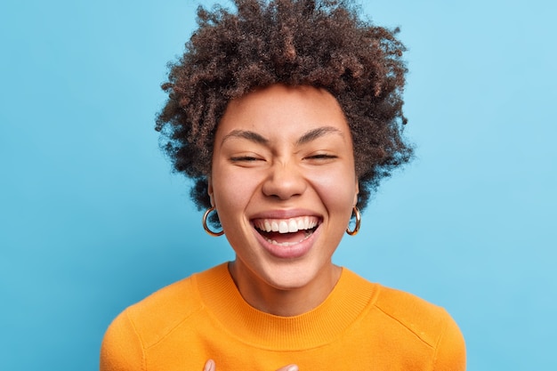 Close up shot of beautiful young African American woman with natural curly hair smiles broadly has positive look  being in good mood wears orange jumper isolated over blue wall