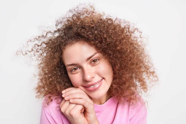 Close up shot of beautiful young female model keeps hands near face tilts head has natural curly hair healthy skin admires someting lovely isolated over white wall. Human face expressions