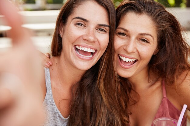 Close up shot of beutiful females with glad looks pose at camera with cheerful expression, pose for selfie