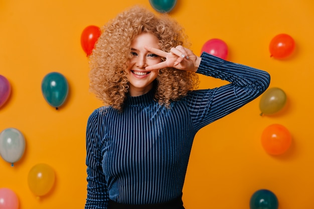 Free photo close-up shot of blue-eyed curly girl in blue striped auitfit showing peace sign on yellow space with multicolored balloons.
