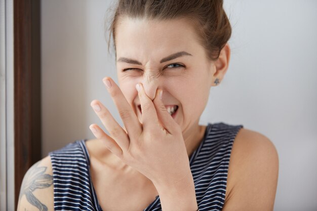 嫌な臭いを避けるために鼻をつまんで、嫌悪感を示す白人の女の子のクローズアップショット。ひどい悪臭への嫌悪感で目を細めている髪の束とブルネットの少女。否定的な感情、厄介な感情。