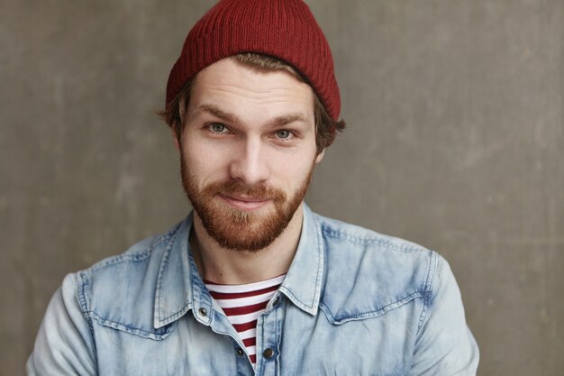 Close up shot of charismatic fashionable young European male with thick beard wearing trendy hat