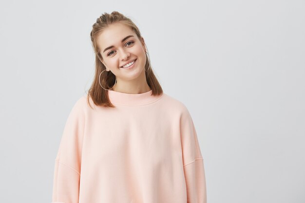 Close up shot of charming Caucasian female with fair straight hair dressed in pink, gently looking , smiling while listening to interesting stories and good news. Tender, beautiful, young.