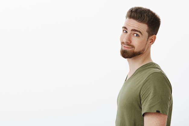 CLose-up shot of charming charismatic bearded boyfriend in t-shirt standing in profile turning head and grinning delighted and carefree posing happy against white wall