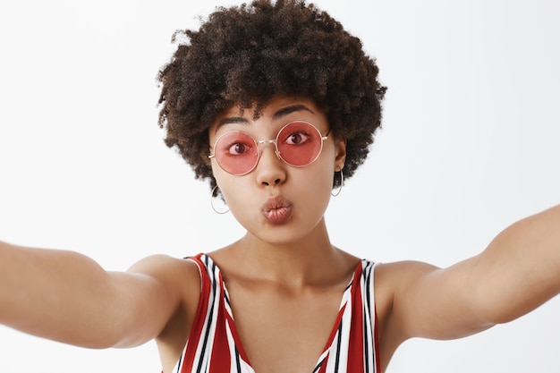 Close-up shot of charming playful and dark-skinned woman with afro hairstyle folding lips in mwah or kiss and pulling hands towards, taking selfie or recording vlog in trendy sunglasses
