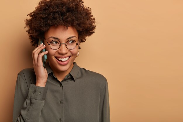 Close up shot of cheerful Afro American woman has hilarious telephone conversation, smiles broadly and looks somewhere pensively