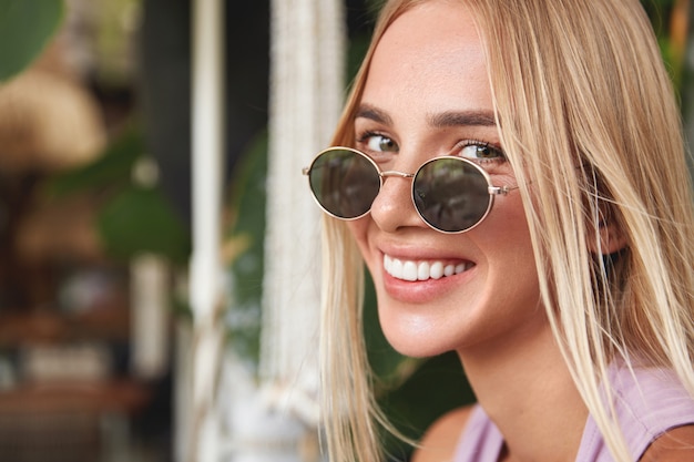 Close up shot of cheerful blonde young woman wears round shades, has broad smile and appealing appearance, being in good mood, looks happily