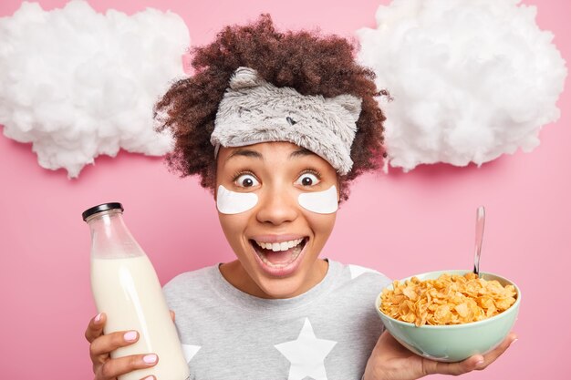 Close up shot of cheerful curly haired Afro American woman looks gladfully at camera applies beauty patches under eyes has breakfast in morning dressed in nightwear isolated over pink wall