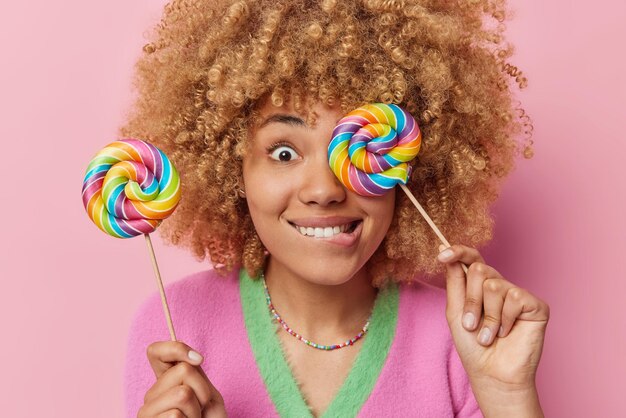 쾌활한 곱슬머리 여성 모델의 클로즈업 샷은 두 개의 맛있는 여러 가지 빛깔의 막대 사탕을 들고 입술을 물고 분홍색 배경 캔디 우먼에 격리된 설탕의 해로운 영향에 관심이 없습니다