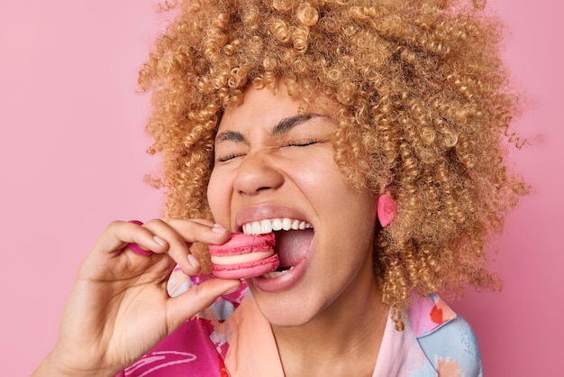 Close up shot of cheerful curly haired woman keeps mouth widely opened bites delicious macaroon enjoys eating sweet food has sugary addiction isolated over pink background. Confectionery concept