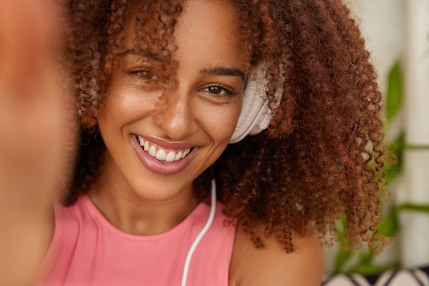 Close up shot of cheerful woman has crisp hair, toothy smile, enjoys recreation time, feels amused, listens favourite music in headphones, makes selfie portrait, models indoor