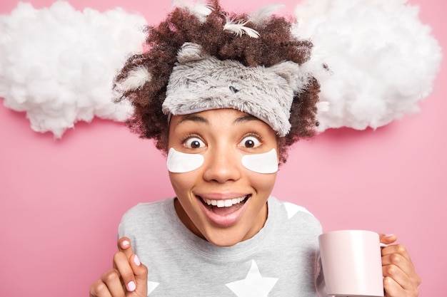 Close up shot of cheerful young woman stares with great surprised at camera drinks refreshing beverage in morning wears sleepmask on forehead slumber suit beauty pads under eyes