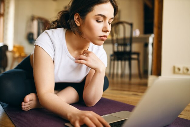 ラップトップを使用してフィットネスマットに座っている集中した若い女性のショットをクローズアップして、ホームトレーニングのオンラインチュートリアルを検索し、ビデオでインストラクターの後にエクササイズを繰り返します。スポーツとガジェット