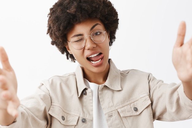 Close-up shot of confident rebel and cool african american lesbian in glasses and beige shirt, showing tongue flirty and winking, pulling hands towards, seducing someone