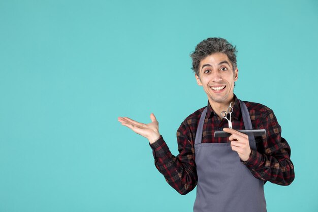 Close up shot of confident young barber wearing gray apron and holding comb and scissor showing something on the right side on blue color background