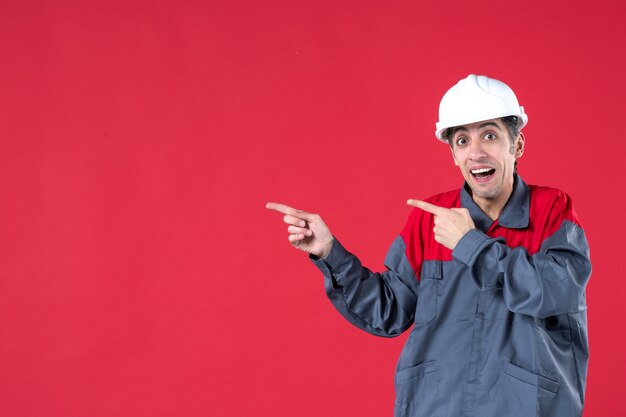 Close up shot of confused young worker in uniform and with hard hat pointing something on the right side on isolated red wall