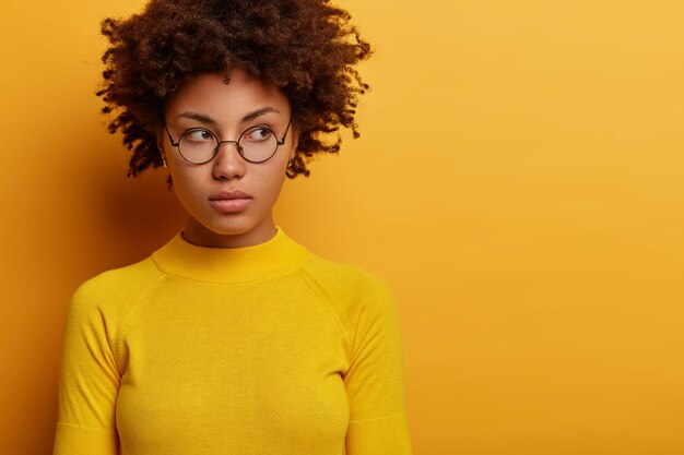 Close up shot of contemplative young female model wears round spectacles ad yellow clothes, looks aside with pensive expression, thinks over plan, poses indoor, blank space for your advertisement