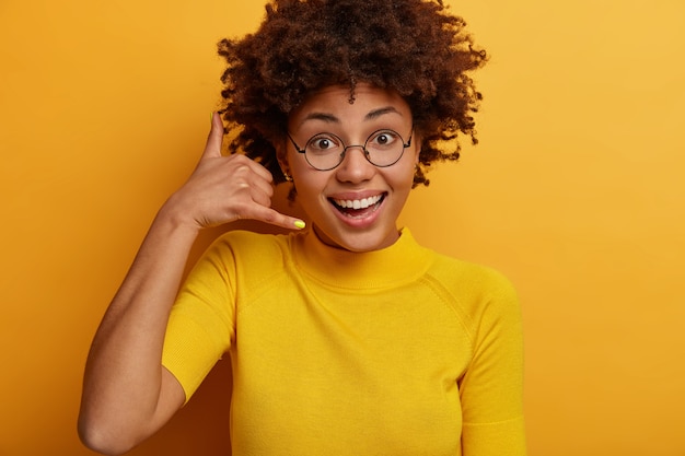 Close up shot of curly beautiful African American woman makes phone gesture, says call me back, wears round glasses and casual t shirt, poses against yellow  wall. Communication sign