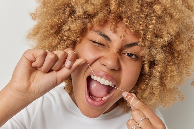 Close up shot of curly haired woman removes food uses thread or dental floss winks eye keeps mouth opened takes care of her mouth cavity poses indoor Teeth flossing and oral hygiene concept