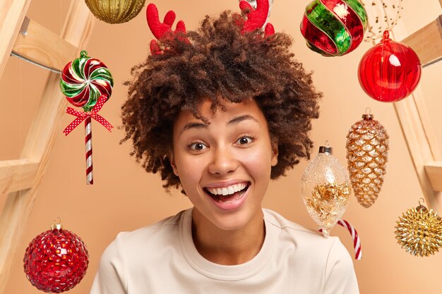Close up shot of curly haired woman smiles broadly has white teeth and healthy dark skin wears red reindeer antlers looks gladfully at camera expresses happiness surrounded by christmas toys
