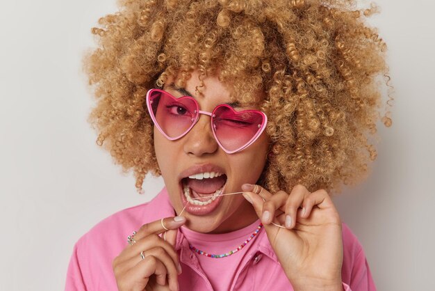 곱슬머리 여성이 윙크하는 눈은 분홍색 선글라스를 착용하고 재킷은 치실을 사용하여 치아를 청소하는 데 사용하여 회색 스튜디오 배경에서 격리된 음식을 제거합니다. 치과 건강 관리 개념