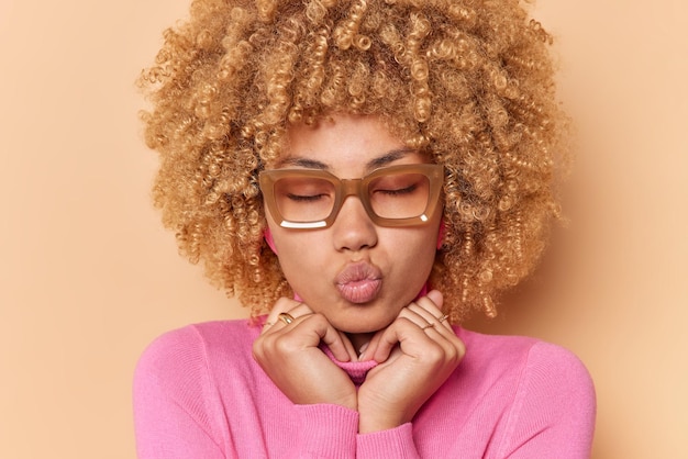 Close up shot of curly haired young woman keeps eyes closed lips folded waits for kiss wears spectacles keeps hands on collar of jumper isolated over beige background Facial expressions concept