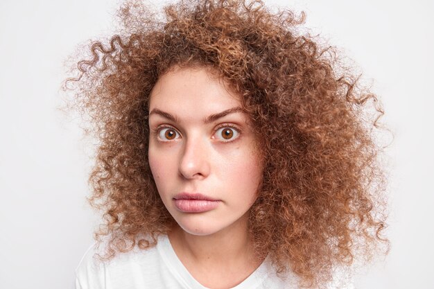 Close up shot of curly wondered young European woman gazes surprisingly  amazes by unexpected news as curly bushy hair isolated over white wall. What did you say is it really so.