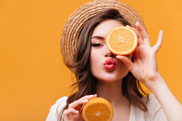 Close-up shot of cute girl in straw hat covering eye with orange and blowing kiss.