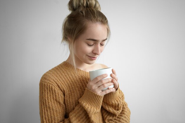 Close up shot of cute pretty girl in cozy knitted sweater enjoying sweet warm cocoa from large cup, closing eyes and inhaling good aroma of hot drink. Beverage, rest, leisure and relaxation