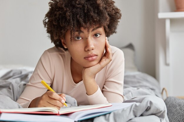 Close up shot of dark skinned girl makes notes of ideas in notebook, lies in bed