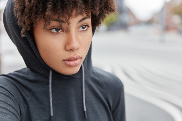 Close up shot of dark skinned hipster has thoughtful expression, wears casual hoodie