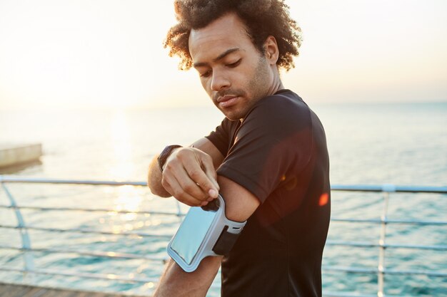 Close-up shot of dark-skinned male athlete fixing mobile arm bag. Morning outdoor workout session behind the sea. Sports, technology and leisure concept.