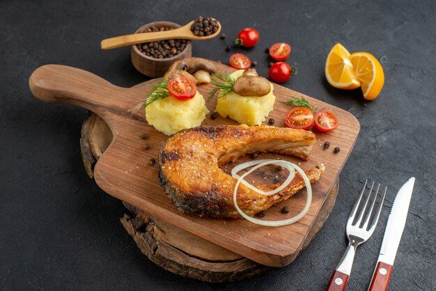 Close up shot of delicious fried fish and mushrooms tomatoes greens on cutting board cutlery set pepper on black surface