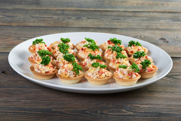 Close up shot of delicious tartlets filled with salad decorated with greens served on white ceramic plate on the wooden table at the local restaurant menu appetizer food tasty.