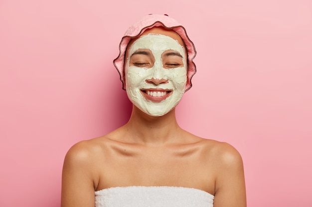 Close up shot of delighted japanese female applies cosmetic face mask, wants to have fresh skin, recreats in spa resort, shows bare shoulders, wears bathcap, has broad smile, isolated on pink wall