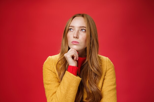 Close-up shot of determined and focused creative thoughtful redhead female looking at upper left corner touching chin, thinking, making choice or remembering information over red background.