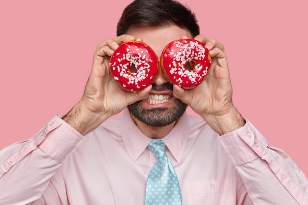 Close up shot of displeased bearded man clenches teeth, covers eyes with doughnuts, dressed formally, stands over pink wall